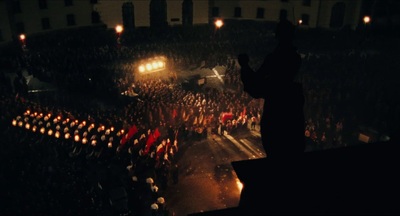 Origin (2023) movie still: wide — A dark outdoor plaza at night filled with a large crowd holding torches and flags. A silhouette of a man stands on…