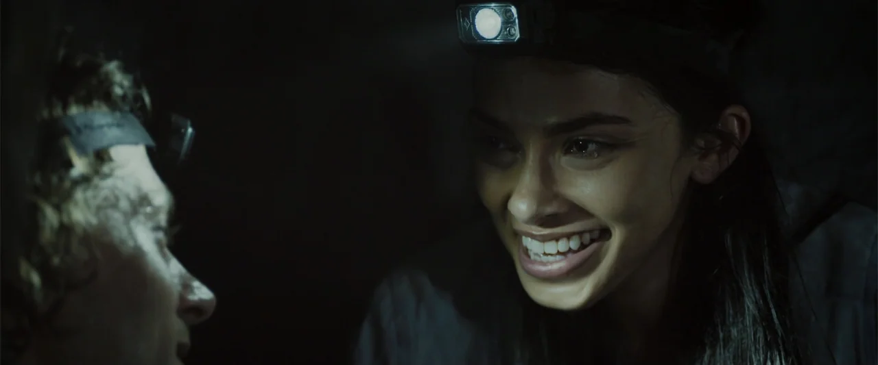 Black Water: Abyss (2020) movie still: medium — A dark, cramped underground cavern environment provides the backdrop for two people. A woman smiles…