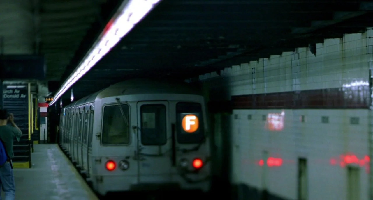 In The Cut (2003) movie still: wide — A subterranean subway platform features white tiled walls and a dark concrete ceiling. An F-line subway train…