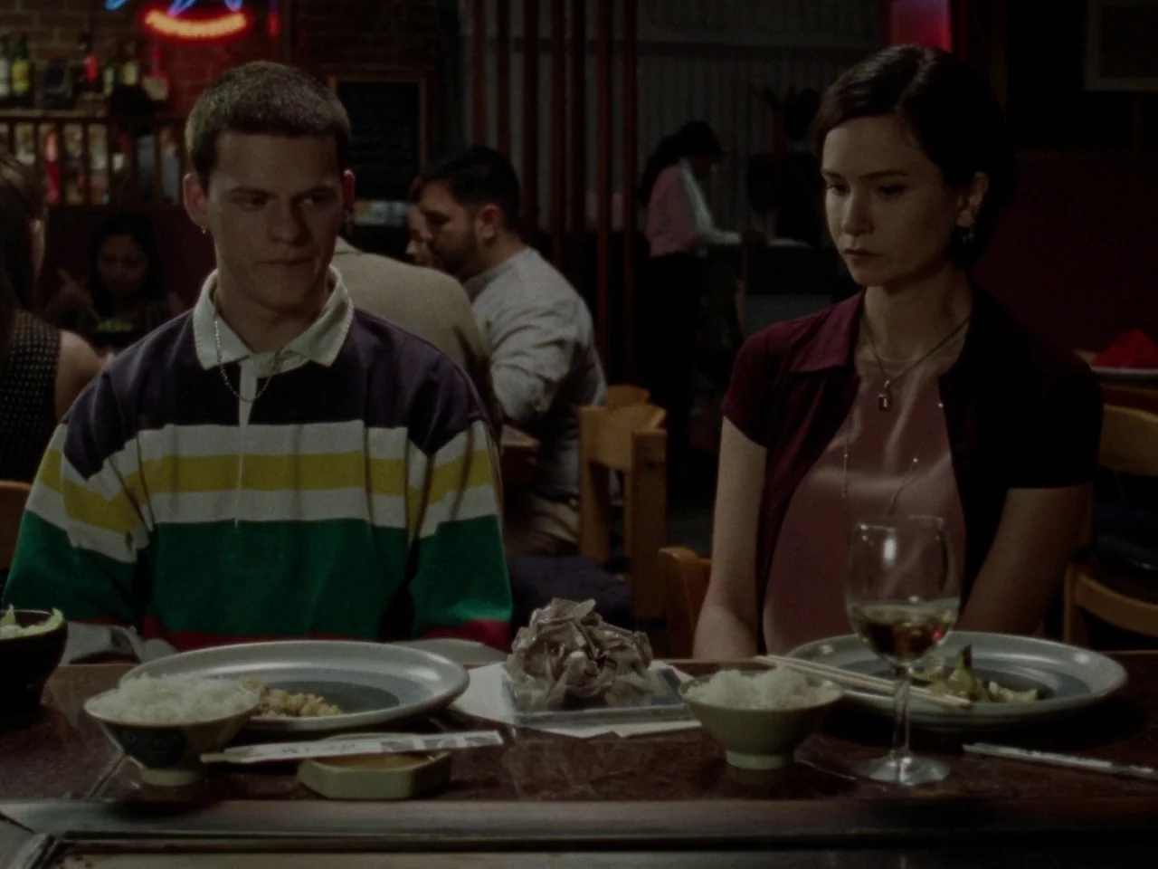 Mid90s (2018) movie still: medium — A dimly lit restaurant setting features wooden tables with plates of rice and side dishes. Two young adults sit…