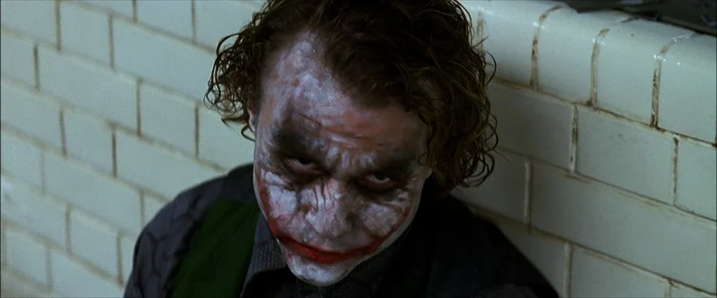 The Dark Knight (2008) movie still: closeup — A man stands against a wall covered in light-colored rectangular ceramic tiles. The character looks…