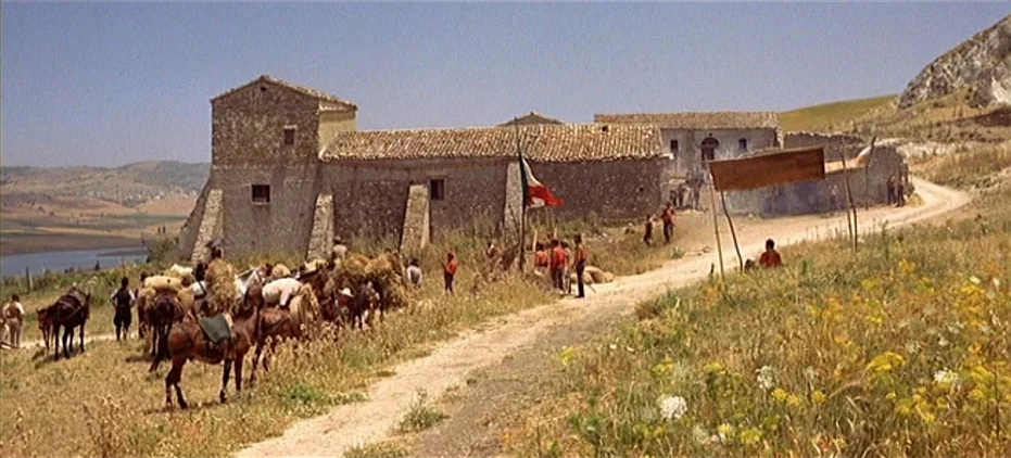 The Leopard (1963) movie still: wide — A stone building with tile roofs sits near a dirt road in arid landscape. People with loaded horses and…