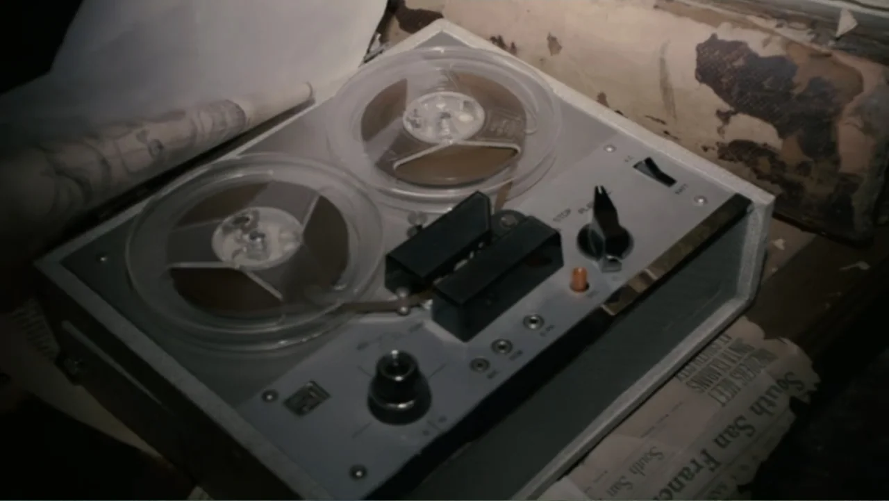 Quarantine (2008) movie still: closeup — A dusty attic surface holds an vintage silver reel-to-reel tape recorder and newspaper. The recorder rests…