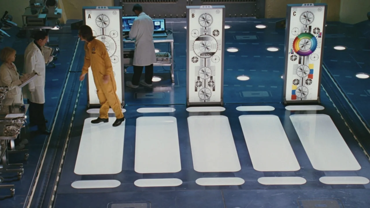 Zoom (2006) movie still: wide — A bright laboratory space features blue flooring and three vertical light-up calibration panels. Three individuals…