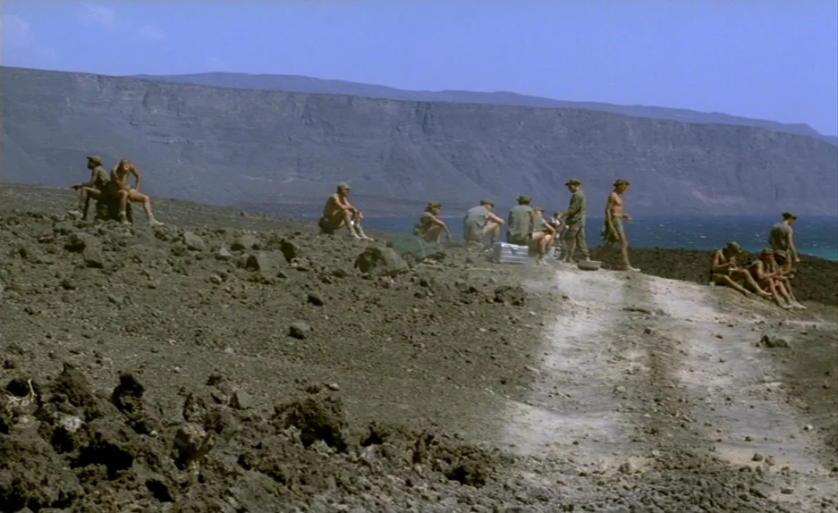 Beau Travail (1999) movie still: wide — A desolate volcanic shoreline features jagged black rocks and a sparse dirt path. Several soldiers in…