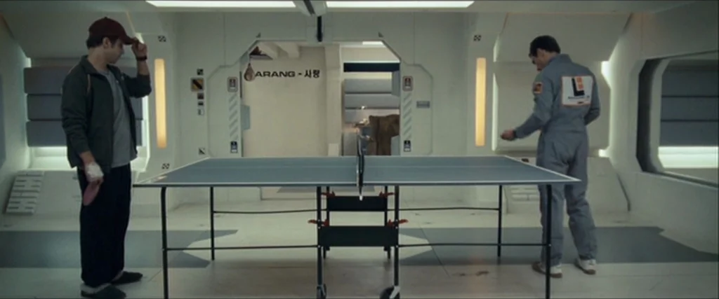 Moon (2009) movie still: wide — A stark white, futuristic interior space resembles a clinical living module or break room. Two men stand facing each…
