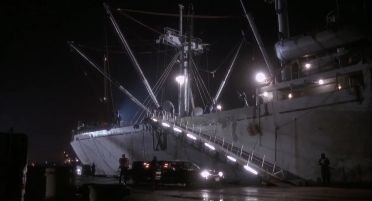 3 Ninjas (1992) movie still: wide — A large cargo ship is docked at a concrete pier at night. Lights illuminate the metal gangplank leading from the…