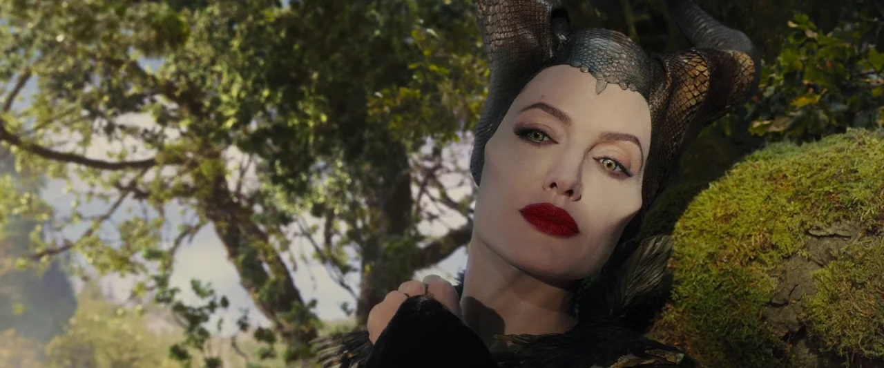 Maleficent (2014) movie still: closeup — A woman with large black horns stands in a sun-dappled deciduous forest. The subject faces the camera while…