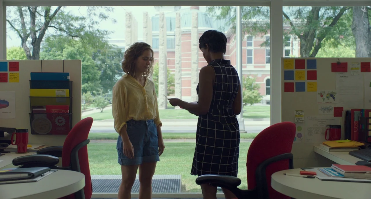 Columbus (2017) movie still: medium — An office interior features two partitioned desks facing a large window with campus views. A woman in a yellow…
