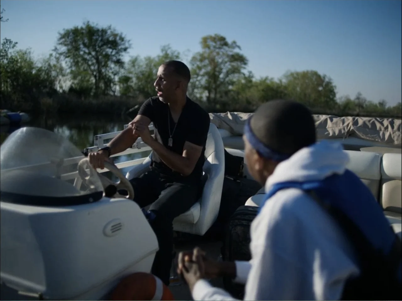 As We Speak: Rap Music on Trial (2024) movie still: medium — A pontoon boat drifts on a calm river surrounded by trees under a clear sky. A man in a…