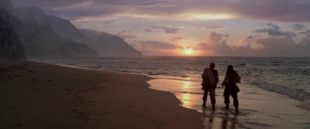 Pirates of the Caribbean: On Stranger Tides movie still: wide — A wide sandy beach stretches toward a rocky cliffside under a golden sunset sky. Two…