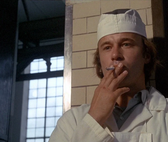 Crime & Punishment (1983) movie still: medium — A man in a work uniform stands against a tiled wall near a window. He holds a lit cigarette to his…