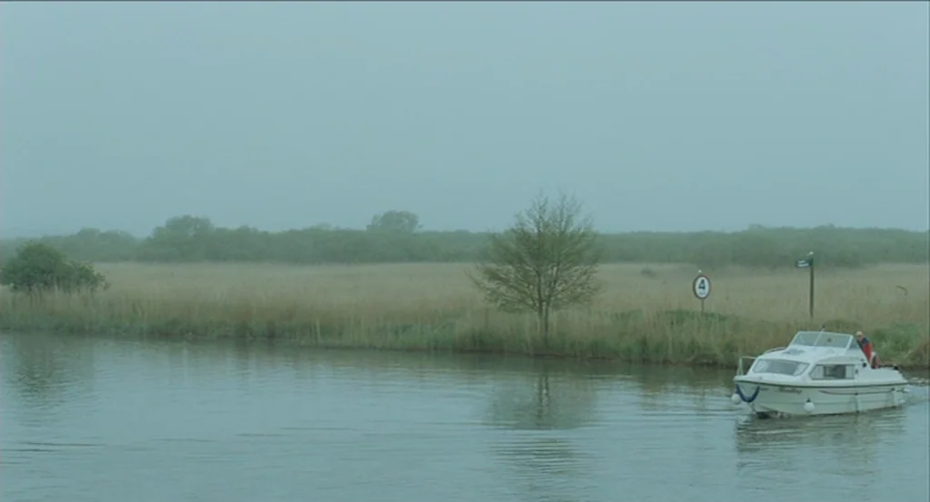 45 Years (2015) movie still: wide — A calm, wide river flows past a flat expanse of tall dry grasses and trees. A small white cabin cruiser boat…