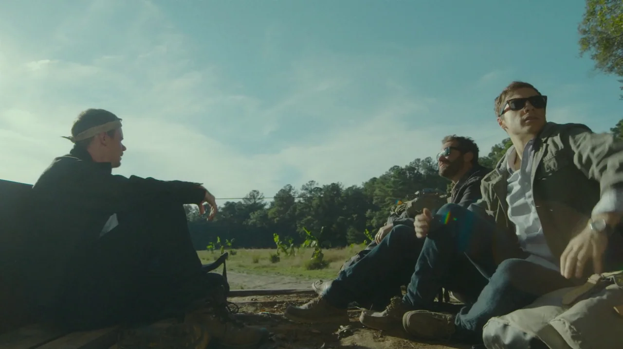 The Sacrament (2013) movie still: wide — Three men are seated in the bed of an open truck in a rural field. They are engaged in conversation while…