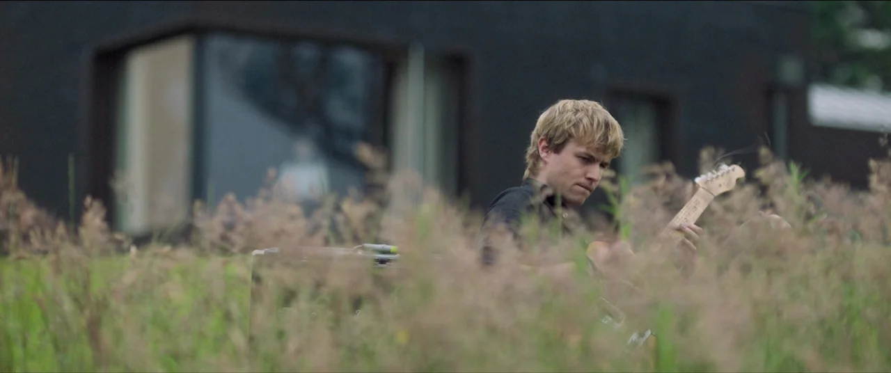 The Feast (2021) movie still: medium — A man plays an electric guitar in a field of tall, unkempt grass. A modern black house with large glass…
