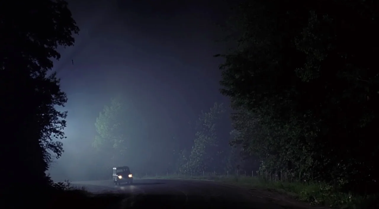 eXistenZ (1999) movie still: wide — A dark, winding road is enveloped in thick, low-lying fog under a night sky. A single small vehicle travels…
