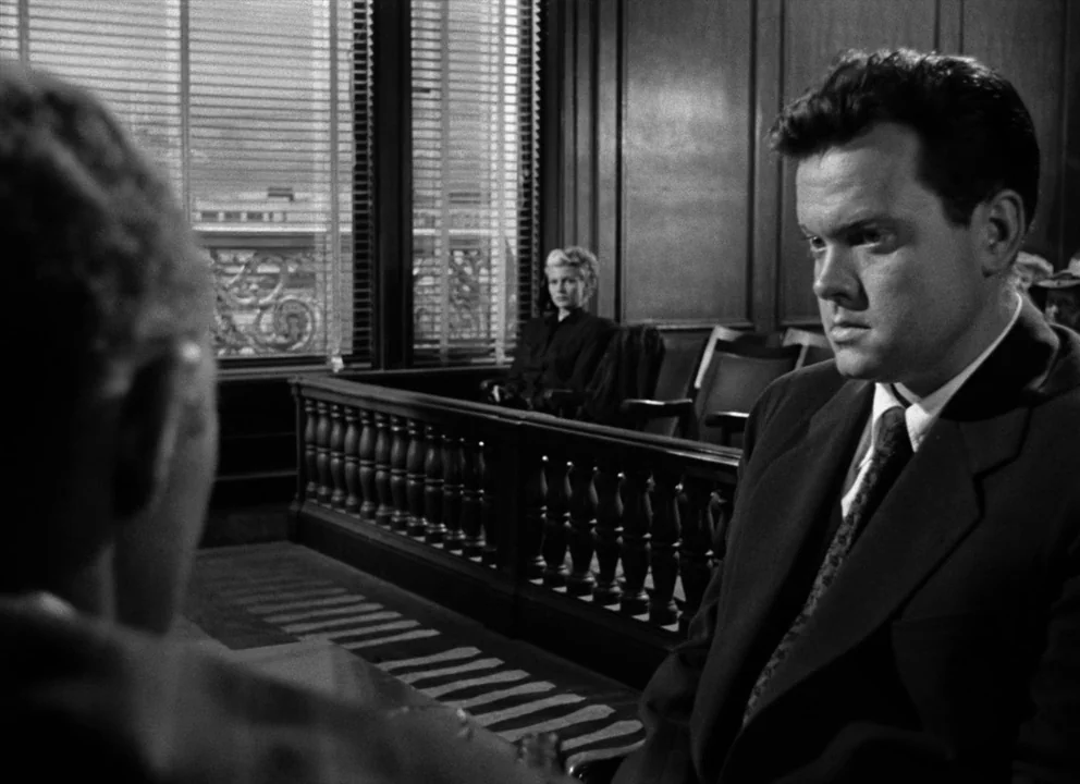 The Lady from Shanghai (1947) movie still: medium — A courtroom interior features dark wood paneling and windows with closed venetian blinds. A man…
