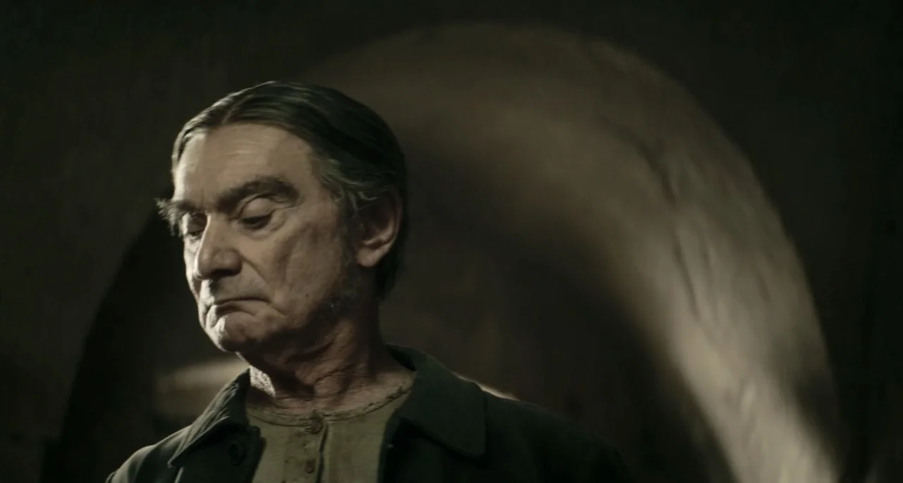 Pinocchio (2022) movie still: closeup — A dark, arched stone interior with rough texture and shadows. An elderly man looks downward while standing…