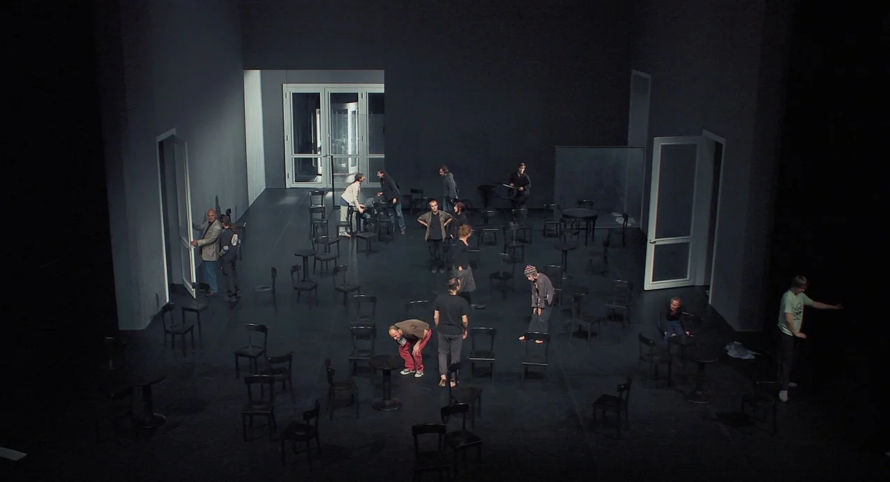 Pina (2011) movie still: wide — A vast, sparsely furnished dark room contains many scattered wooden chairs on a flat floor. Several individuals…