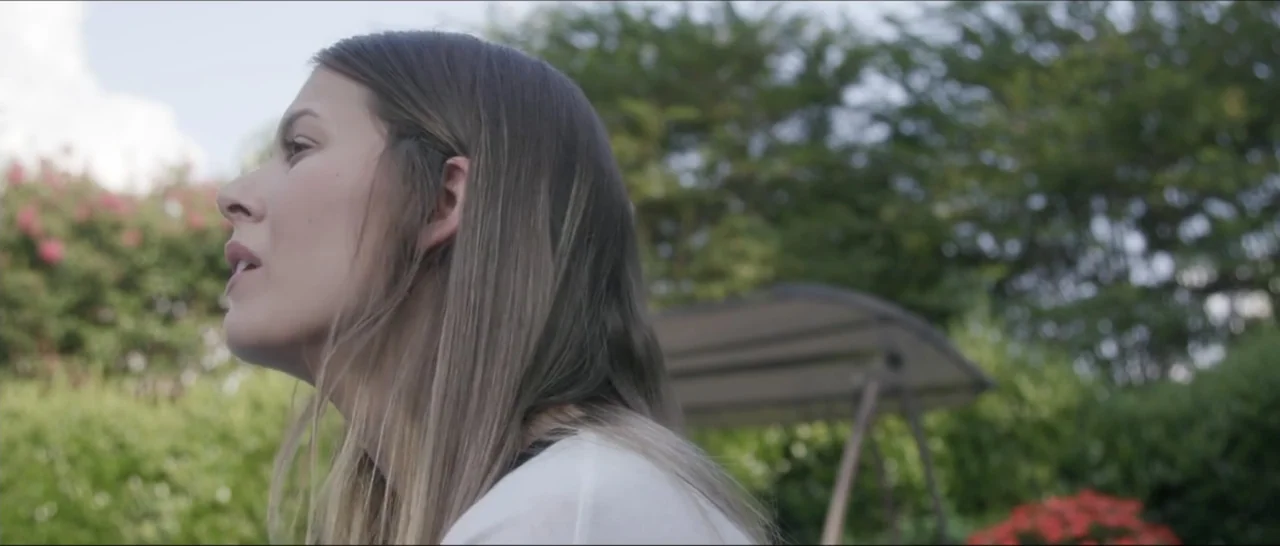 If You're Gone (2019) movie still: medium — A woman stands outdoors in a green backyard filled with flowering shrubs and trees. She is captured in…