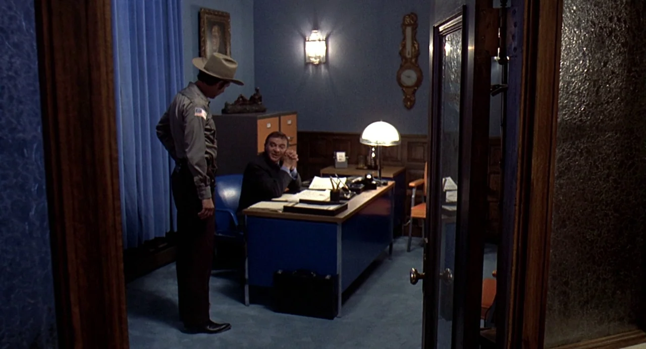 Charley Varrick (1973) movie still: wide — A 1970s professional space featuring blue carpet, wood-paneled walls, and a large metal desk. An officer…