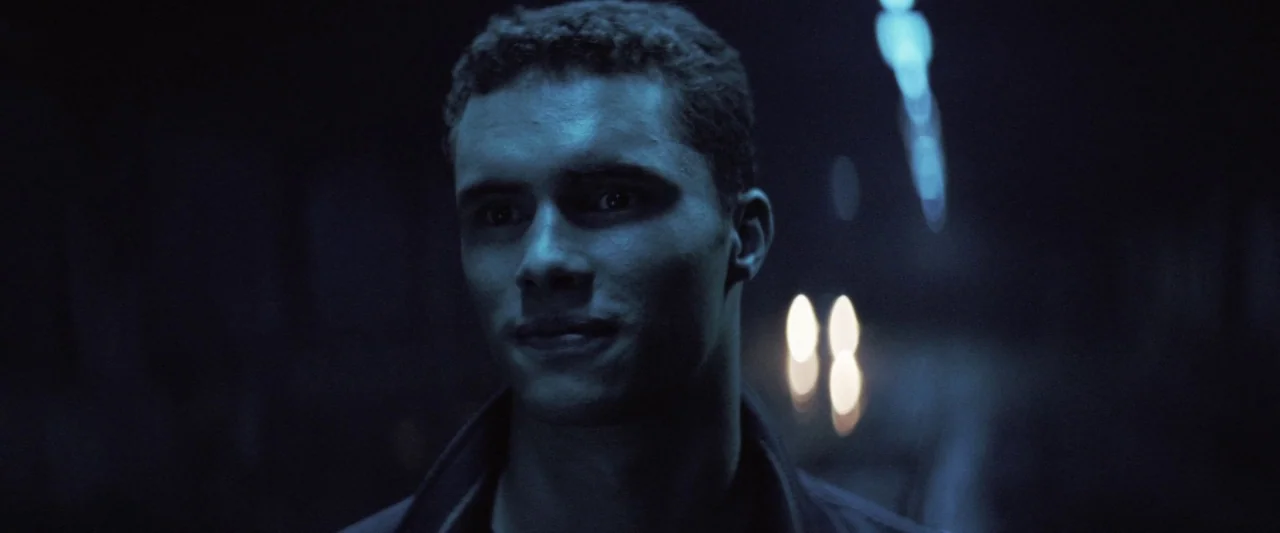 Colt 45 (2014) movie still: closeup — A dark outdoor street at night with blurred lights in the background distance. A young man with short curly…