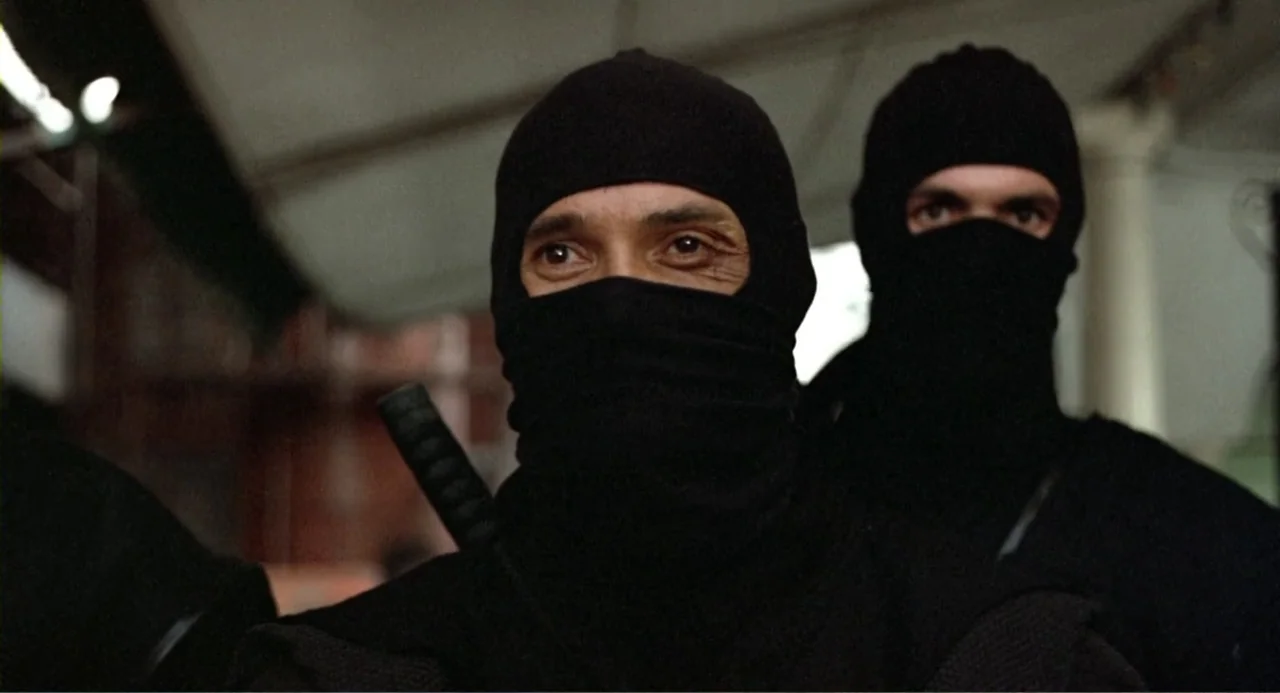 American Ninja 2: The Confrontation (1987) movie still: medium — Two individuals wearing black ninja-style head coverings and shirts occupy an urban…