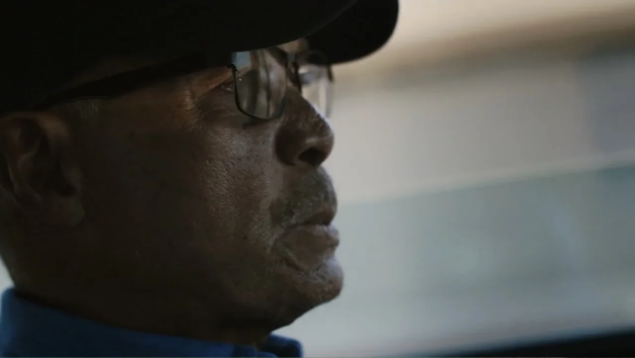Reggie (2023) movie still: closeup — The interior of a vehicle during daylight hours with motion blur visible outside. An older black man in a blue…