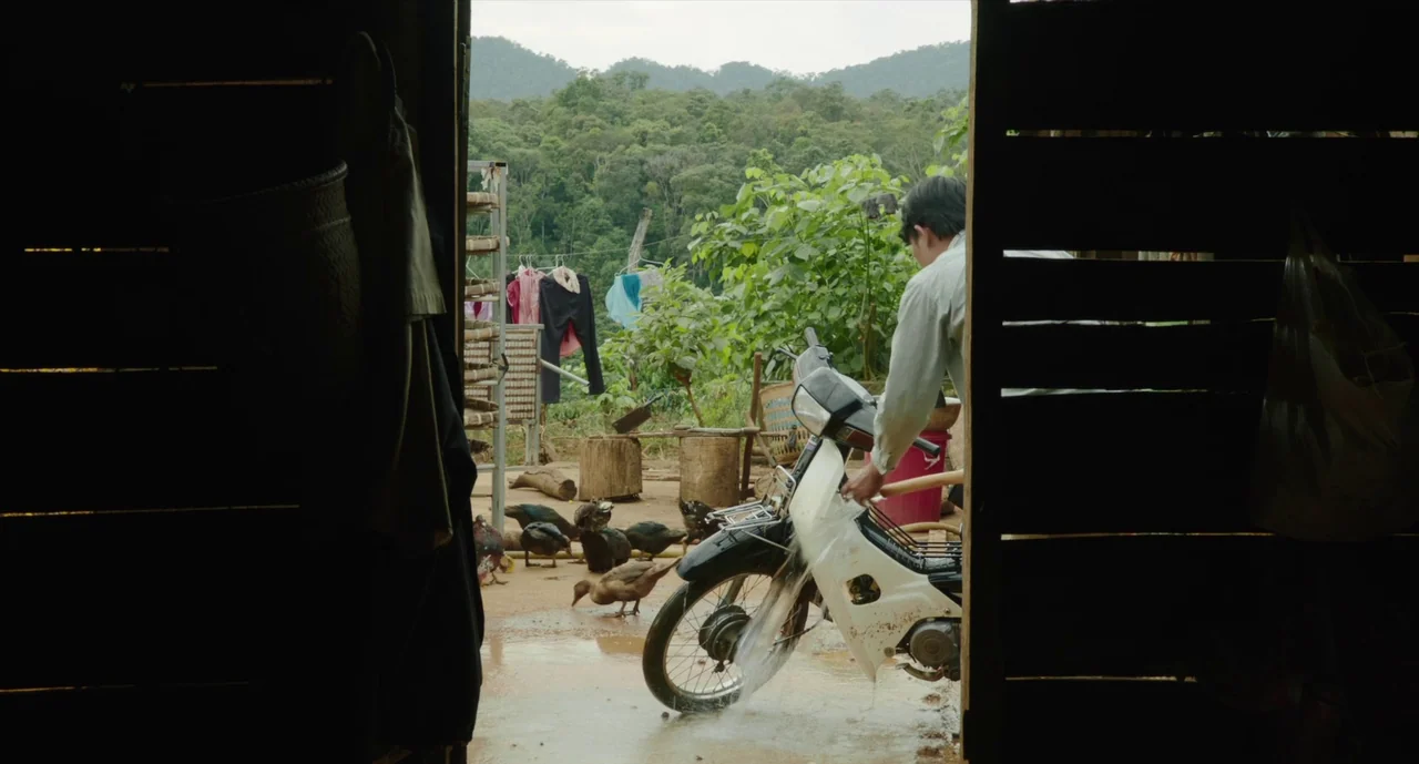 Inside The Yellow Cocoon Shell (2023) movie still: medium — An outdoor farmyard view framed from within a dark, wooden-walled structure. A man…