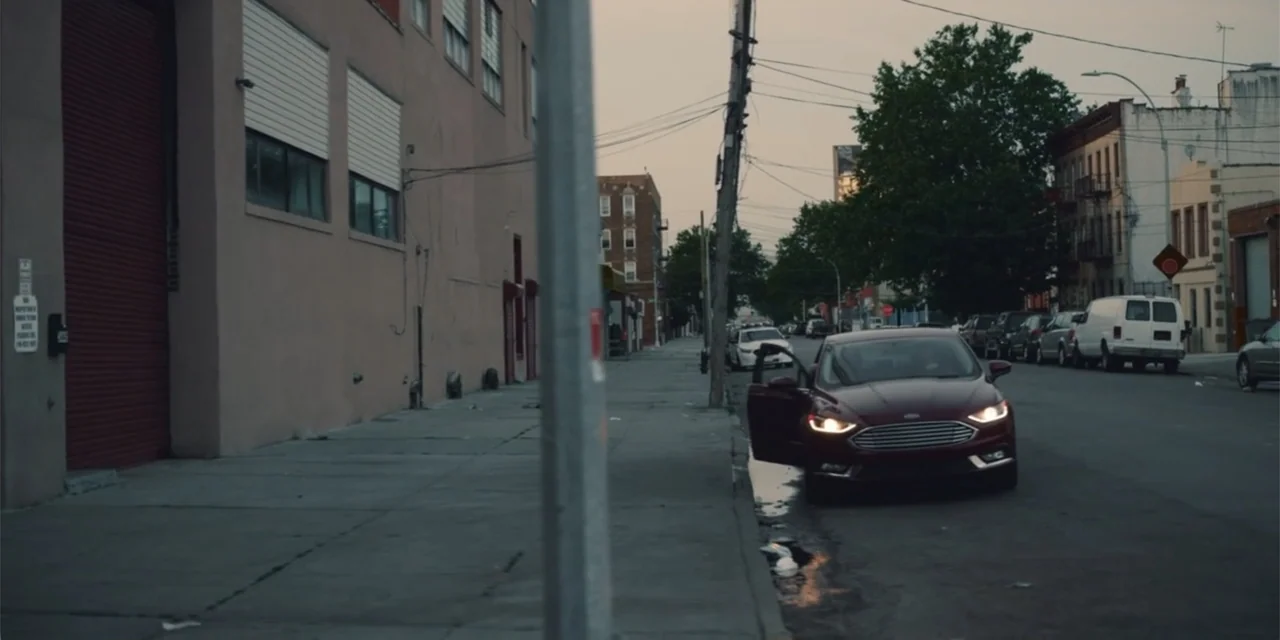 Equal Standard (2021) movie still: wide — An urban street at dusk features concrete buildings and a parked sedan with doors open. A red car idles at…