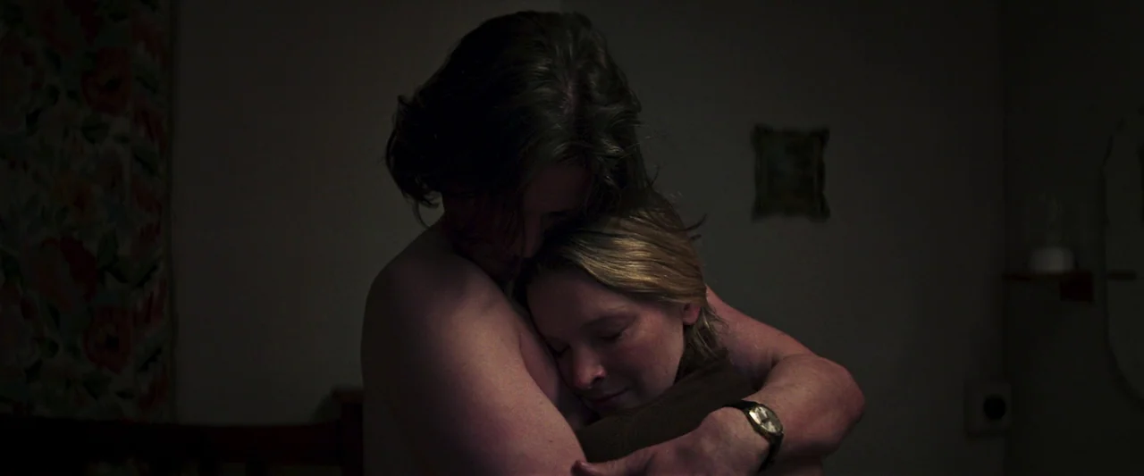Starve Acre (2023) movie still: medium — A dimly lit room with plain walls and a small picture frame in the background. A man holds a blonde woman…