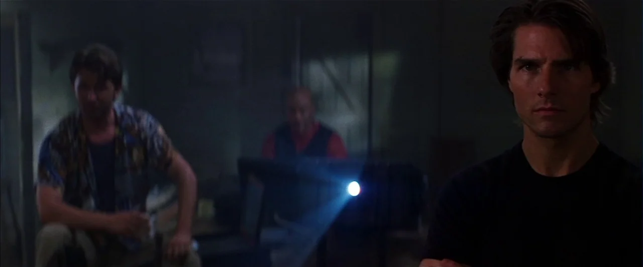 Mission: Impossible II (2000) movie still: medium — A dimly lit room with industrial features contains three men watching a projection. The…