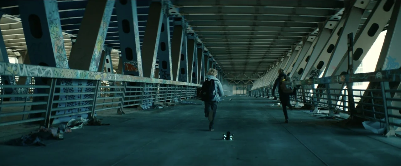 Invasion (2020) movie still: wide — A long, graffiti-covered concrete pedestrian path runs beneath the massive steel girders of a bridge. Two…