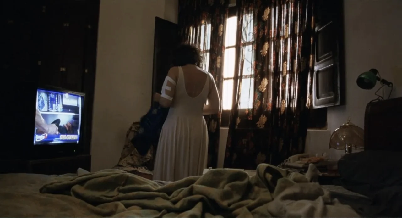 La Ciénaga (2001) movie still: fullbody — A dark hotel room with patterned curtains and an unmade bed in the foreground. A woman faces the window…