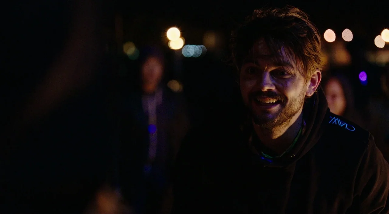 Urge (2016) movie still: medium — A night scene on an urban street with blurred city lights in the background. A man with disheveled hair faces…