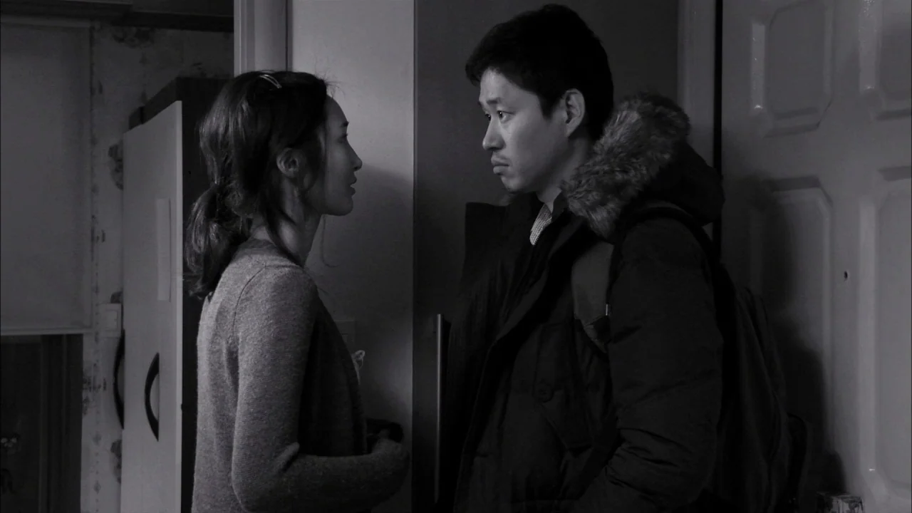 The Day He Arrives (2018) movie still: medium — A dark apartment entryway features a partially visible door frame and paneled door. A man wearing a…