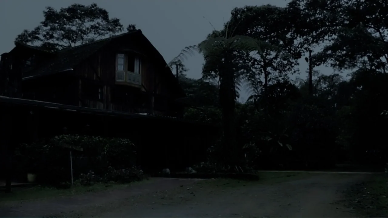 El Duende Sátiro (2016) movie still: establishing — A dark wooden two-story house stands nestled among dense trees at night. The structure is…
