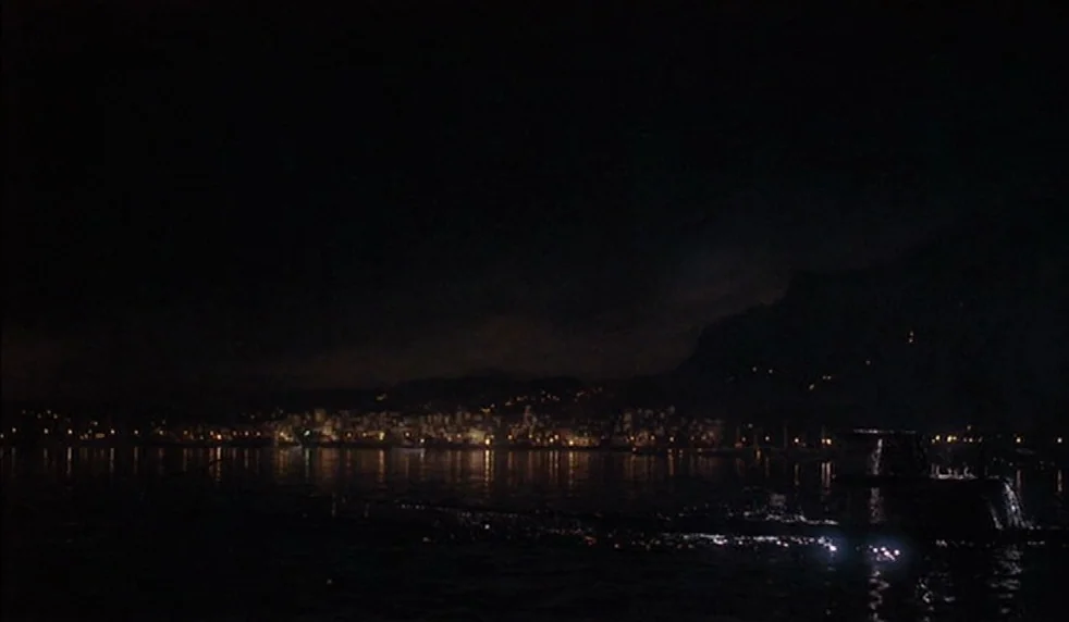 Das Boot (1981) movie still: establishing — A dark harbor at night showing a distant illuminated coastal city against mountains. A submarine conning…