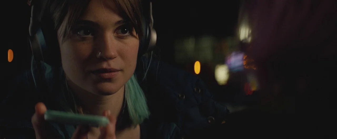 Poser (2021) movie still: closeup — A night scene on an urban street with bokeh lights visible in the background. A young woman with teal-tipped…