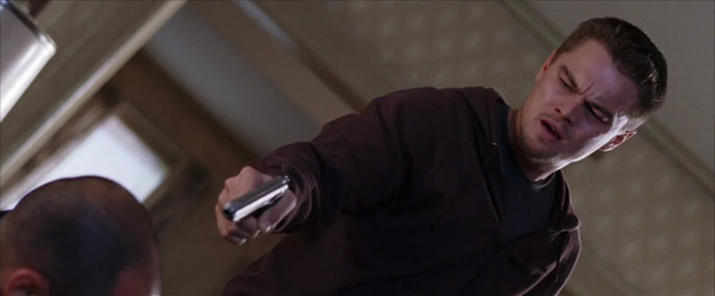 The Departed (2006) movie still: medium — A dimly lit room with muted-colored wallpaper and simple domestic furnishings. A man wearing a maroon…