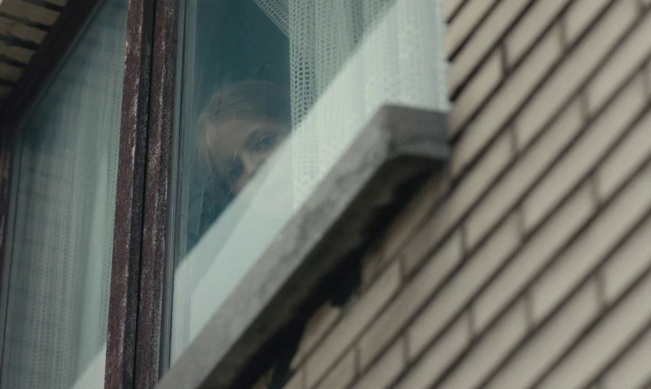 Maldoror (2024) movie still: closeup — A brick building exterior shows a window with a sheer white curtain inside. A young person looks out from…