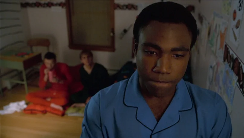 Mystery Team (2009) movie still: closeup — A dimly lit room with light-colored walls and posters attached to the side wall. A young man in blue…