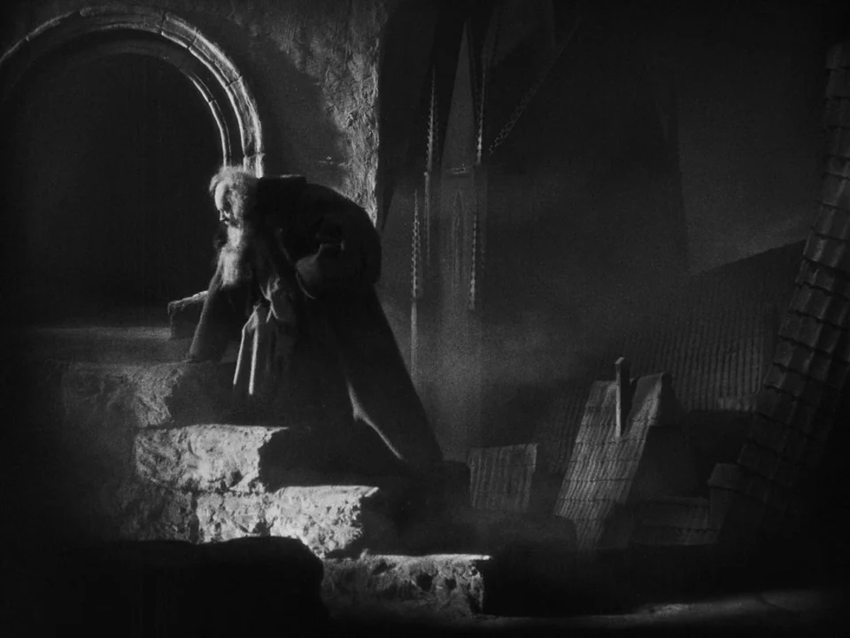 Faust (1926) movie still: fullbody — A dark, stone architectural space with heavy masonry and arched passageways filled with fog. An elderly man…