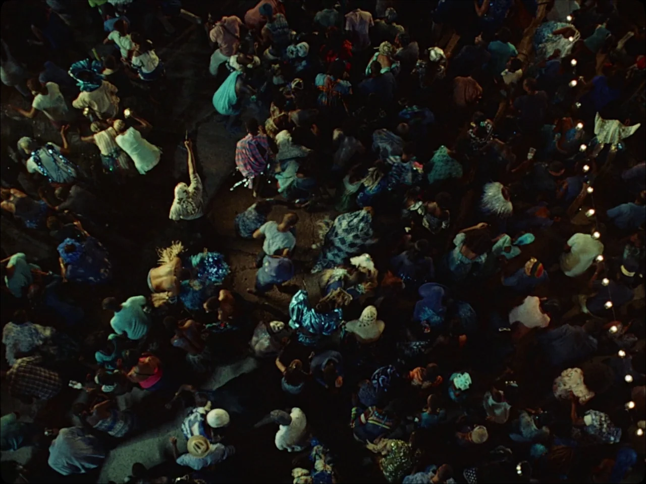 Guava Island (2019) movie still: establishing — An aerial view captures a large, tightly packed crowd gathered for an evening event. Rows of small…