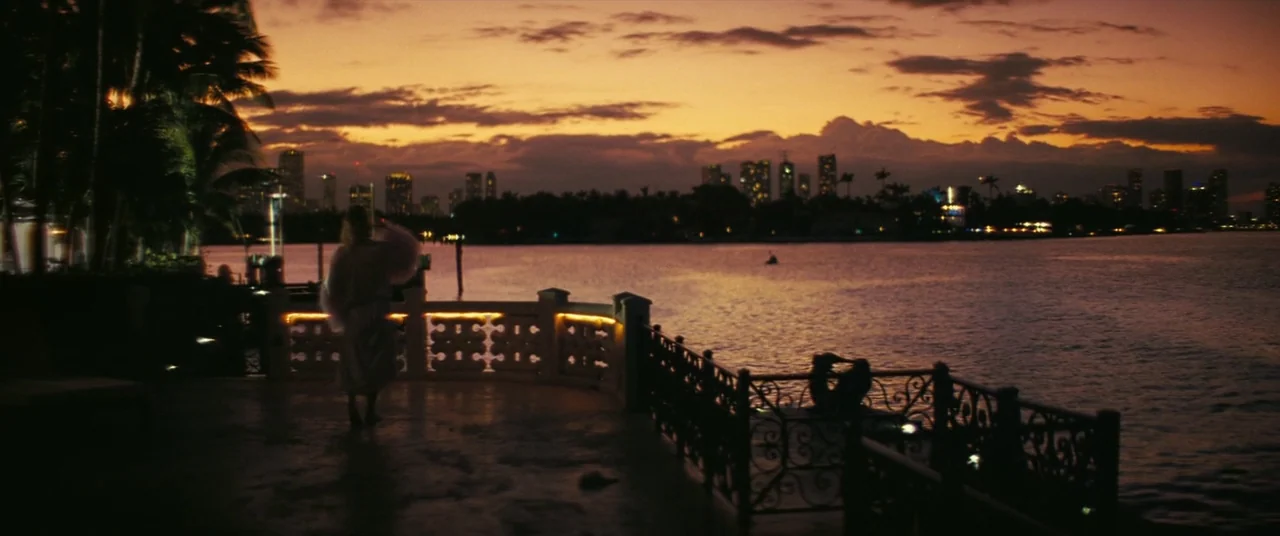 The Beach Bum (2019) movie still: wide — A waterfront balcony at dusk features decorative railings and silhouettes of tropical trees. A lone figure…