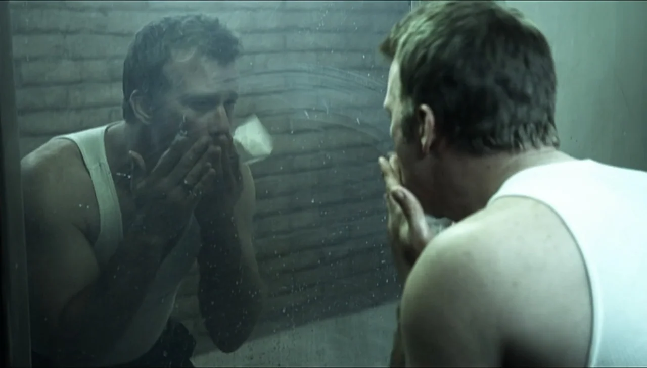 Dark Country (2009) movie still: medium — A dimly lit industrial style space with brick walls viewed through a dirty glass mirror. A man faces his…