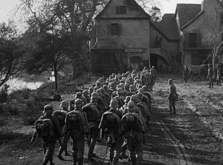 All Quiet on the Western Front (1930) movie still: wide — A column of soldiers marches along a muddy road near aged European stone houses. A single…