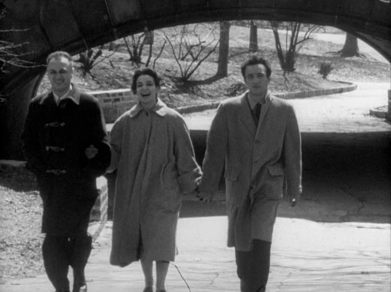 Shadows (1958) movie still: fullbody — A stone bridge provides shelter as three people walk together along a paved walkway. The surrounding park…