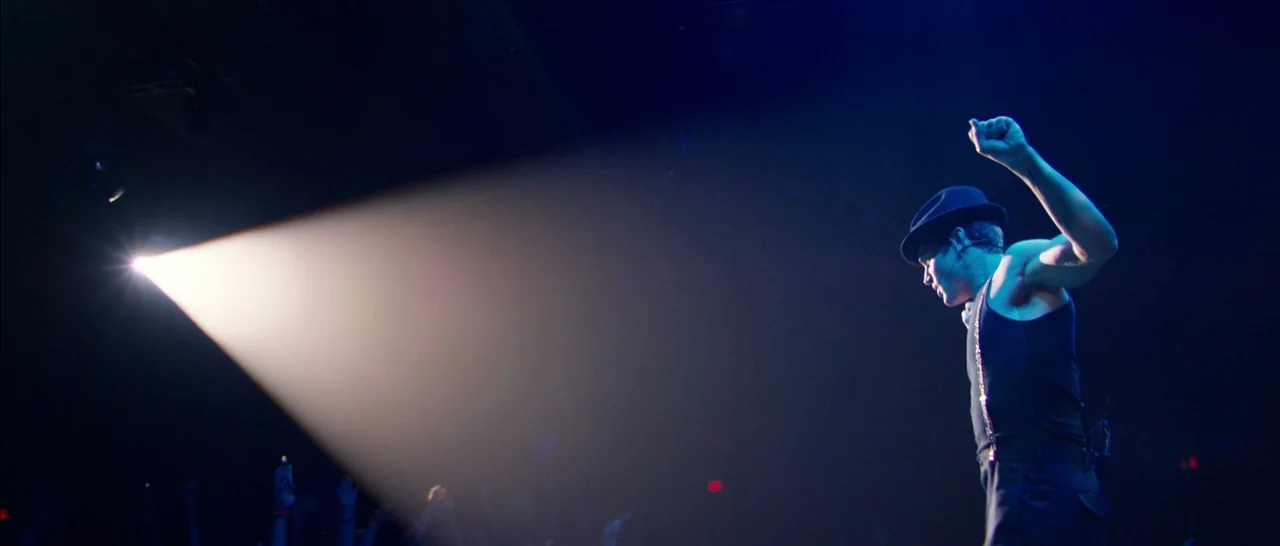 Magic Mike XXL (2015) movie still: medium — A dark concert venue with a crowd faintly visible at the bottom of frame. A male performer wearing a…