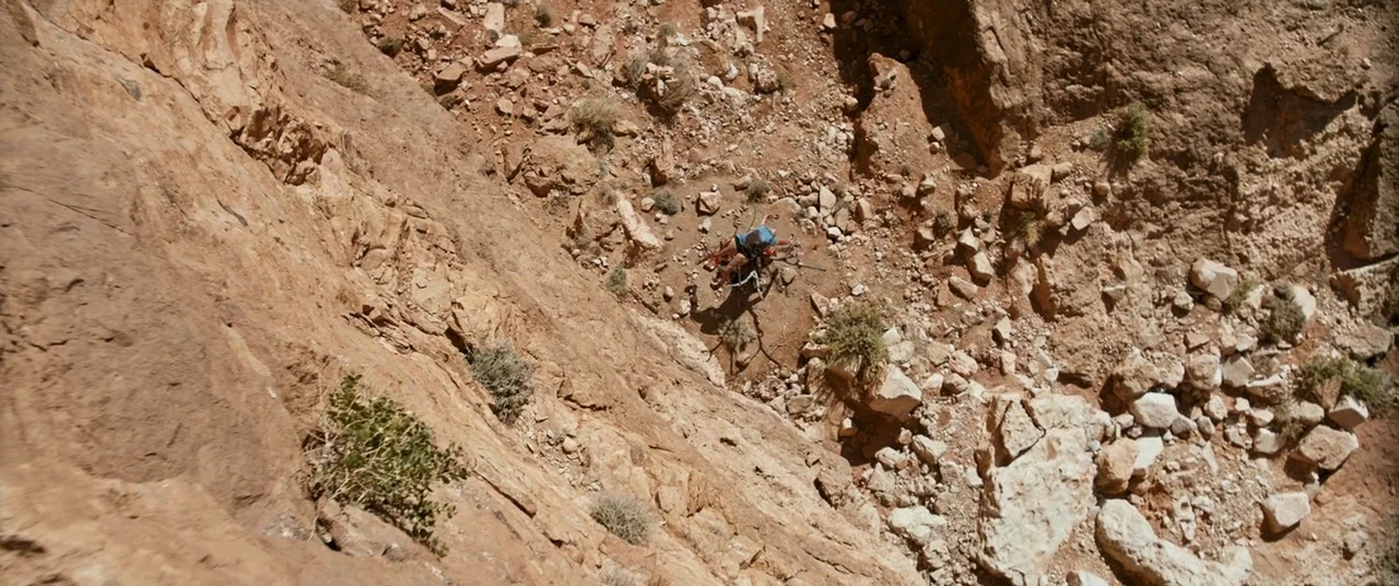 Revenge (2017) movie still: wide — A steep-walled, rugged desert canyon floor is covered in loose rocks and sparse scrub. A lone person lies…